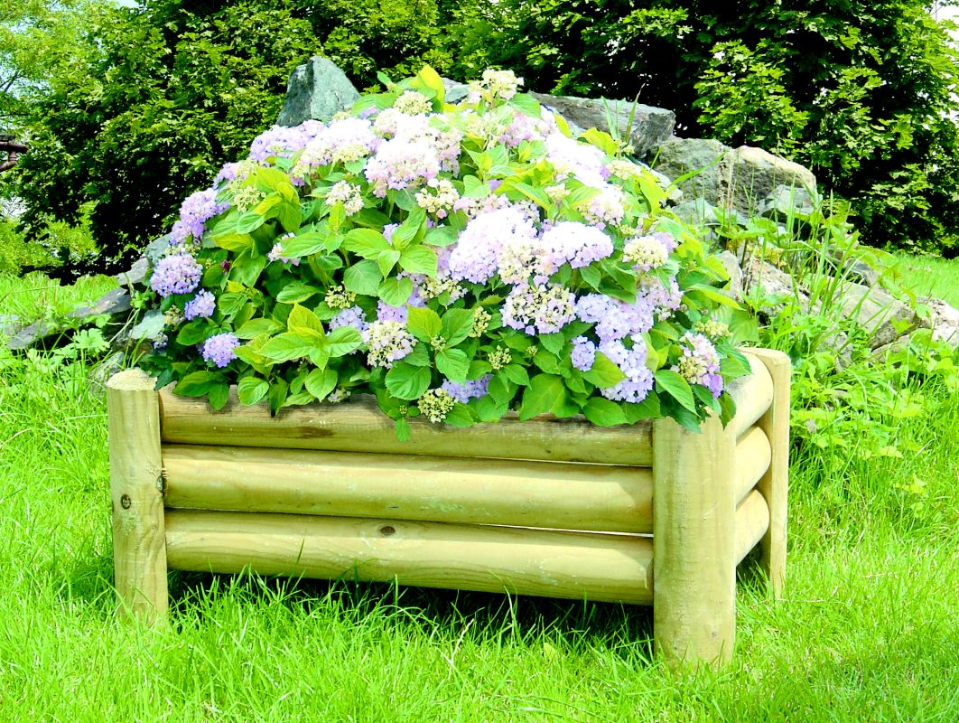 Drewniana donica ogrodowa wypełniona kwitnącymi hortensjami w odcieniach fioletu i bieli, ustawiona na trawniku przed zielonymi krzewami i skałami.